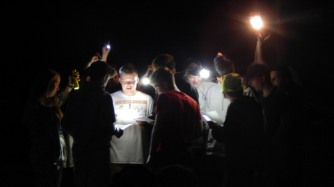 cabin group gathered together in the dark with flashlights to help them sing lyrics written on papers they are holding.