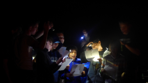 group of boys gathered together in the dark with flashlights to help them sing lyrics written on papers they are holding.