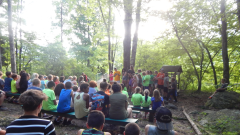 campers seated on benches in a wooded area watching a performance by a platoon group in front.