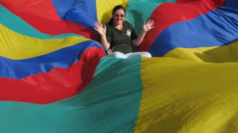 Counselor smiles from the wavy center of a colorful parachute.