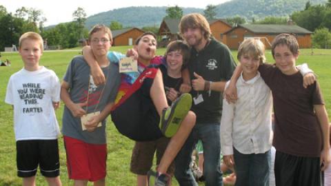 group of smiling boys stand with their cabin counselor. One boy is held off the ground by two others.