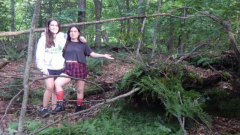 Two youth in front of a shelter made with sticks and ferns. 