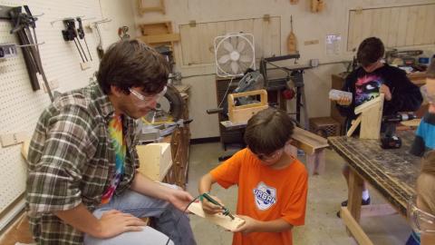 counselor helps camper use wood working tool in camp wood shop 
